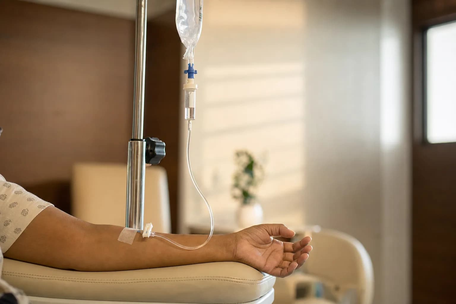 A patient's relaxed forearm receiving a clear IV infusion in a calm treatment suite - the IV bag, drip chamber, and cannula in soft afternoon light